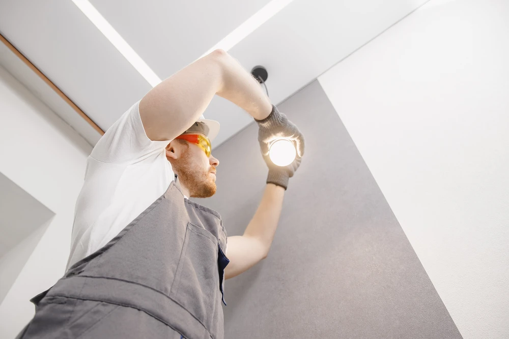 Électricien professionnel en salopette grise et t-shirt blanc installe un luminaire encastré au plafond dans un intérieur moderne