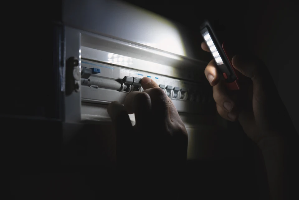 Électricien professionnel en équipement de sécurité (gilet, chaussures de sécurité) intervenant chez un client dans un intérieur residential, effectuant un diagnostic ou une réparation sur un disjoncteur ou un tableau électrique. Le client observe à distance, confiant. L'ambiance générale est professionnelle, méthodique et rassurante, illustrant la compétence technique et le respect des normes de sécurité.