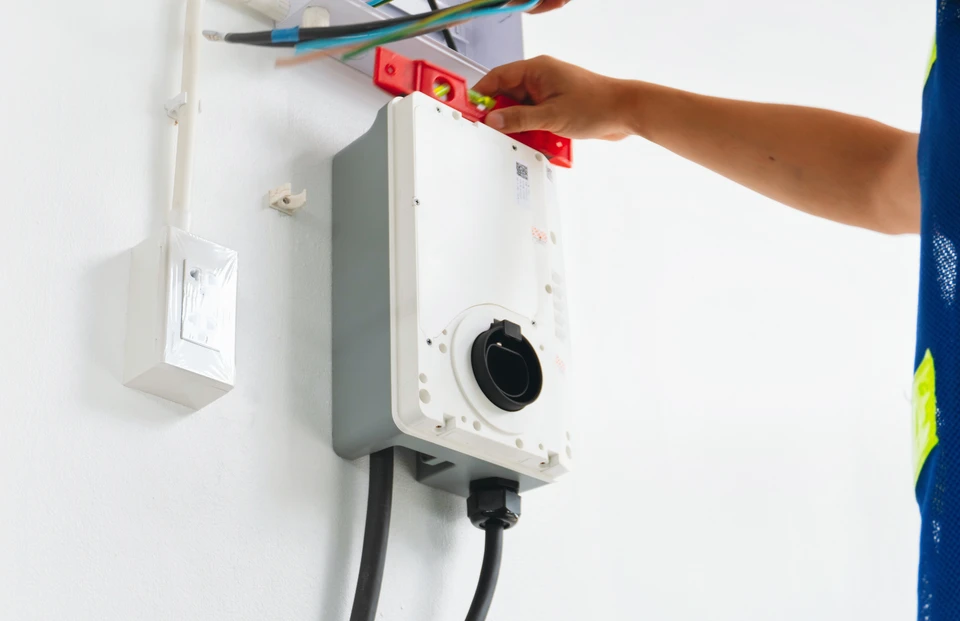Photographie r&eacute;aliste d'un &eacute;lectricien professionnel effectuant l'installation d'une borne de recharge pour v&eacute;hicule &eacute;lectrique sur la fa&ccedil;ade ext&eacute;rieure d'une maison r&eacute;sidentielle ou d'un immeuble tertiaire. L'&eacute;lectricien porte des &eacute;quipements de s&eacute;curit&eacute; (gilet, outils professionnels), travaille m&eacute;thodiquement sur le c&acirc;blage et le branchement de la borne. Le contexte montre clairement une installation compl&egrave;te et soign&eacute;e, repr&eacute;sentative de services de mobilit&eacute; &eacute;lectrique professionnels et certifi&eacute;s IRVE.